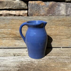 May include: A small, blue ceramic pitcher with a rounded body and a handle. The pitcher is sitting on a wooden surface.