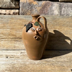 May include: A brown ceramic pitcher with a whimsical face and painted grapes. The pitcher has a handle and a wide mouth.