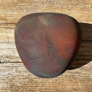 May include: A dark brown ceramic vase with a rounded, heart-shaped design. The vase has a mottled, textured surface with hints of red, blue, and green.