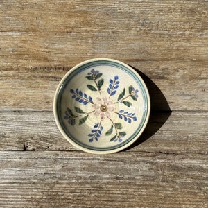 May include: A small, white ceramic bowl with a blue and green floral design. The bowl has a white background with blue and green flowers and leaves.
