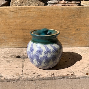 May include: A ceramic jar with a lid, featuring a white and purple speckled glaze. The lid is a dark teal green color.