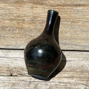 May include: A dark green ceramic vase with a rounded body and a narrow neck. The vase has a glossy finish and is sitting on a wooden surface.