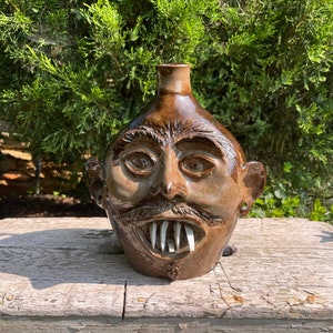 May include: A brown ceramic jug shaped like a face with a wide mouth and large teeth. The jug has a mustache and eyebrows.