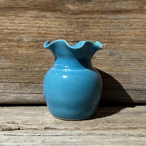 May include: A small, turquoise ceramic vase with a ruffled rim. The vase has a rounded body and a slightly flared neck. The glaze is smooth and glossy, reflecting the light. The vase is displayed on a wooden surface.