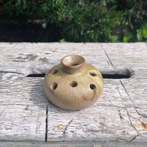 May include: A small, round, brown and green ceramic bud vase with multiple holes around the top for flower stems.