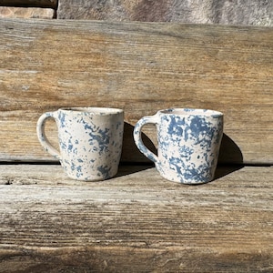 May include: Two ceramic mugs with a white base and blue speckled glaze. The mugs have a rounded shape and a handle on one side.