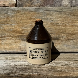 May include: A vintage ceramic jug with a brown top and a beige base. The jug has a cork stopper and a rectangular label that reads "HIGH GRADE SHERRY WINE SALZMAN & SIEGELMAN BROOKLYN, N. Y."
