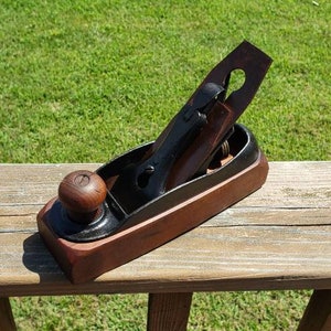 May include: A vintage wooden hand plane with a dark finish, featuring a black metal blade and a wooden knob. The tool is set against a natural wood surface with a green grassy background.