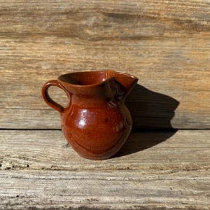 May include: A small, brown ceramic pitcher with a rounded body and a flared lip. The pitcher has a handle and a glossy finish.