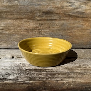 May include: A round, mustard-yellow ceramic bowl with a ribbed exterior. The bowl sits on a wooden surface. The bowl's interior is smooth, and the exterior has concentric ridges. This bowl is suitable for serving food.