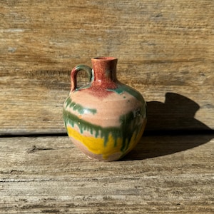 May include: A small, handcrafted ceramic jug with a handle. The jug features a rounded body and a narrow neck. It is painted with a blend of colors, including pink, green, yellow, and red. The jug is displayed on a wooden surface.