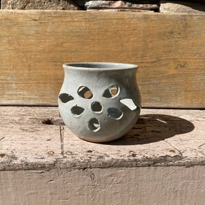 May include: A white ceramic candle holder with a cut-out design. The holder has a round shape and a slightly flared rim.