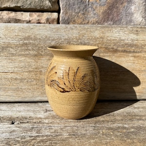 May include: A tan-colored ceramic vase with a wide base and flared rim. The vase features an etched design of tall grass or reeds around its midsection. The vase is set against a weathered wooden surface and a stone background.