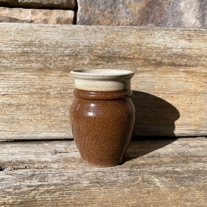 May include: A small, brown ceramic vase with a white rim. The vase has a speckled brown glaze and a rounded body.