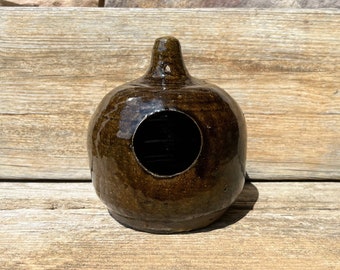 Puede incluir: Una casa para pájaros de cerámica marrón oscuro con una abertura redonda. La casa para pájaros está sobre una superficie de madera.