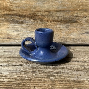 May include: A blue ceramic candle holder with a handle. The holder has a small, round base and a cup-shaped top for holding a candle.
