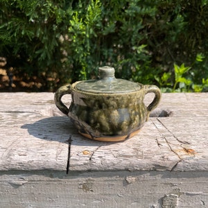 May include: A small, handmade ceramic sugar bowl with a lid. The bowl is a mottled green color with two small handles and a matching lid. The base of the bowl is a light tan color. The bowl is sitting on a weathered wooden surface.