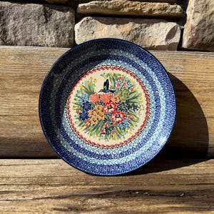 May include: A blue and white ceramic plate with a floral design featuring a hummingbird and colorful flowers. The plate has a wide rim with concentric circles of blue and white.