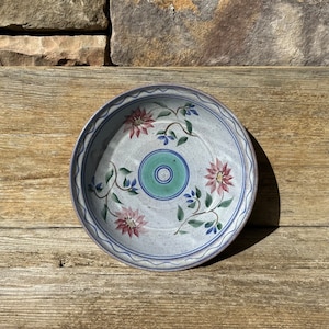 May include: A white ceramic plate with a blue rim and floral design. The plate features three pink flowers with green leaves and a blue and green circle in the center.
