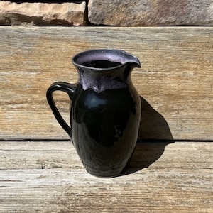 May include: A black ceramic pitcher with a purple rim. The pitcher has a wide mouth and a handle.