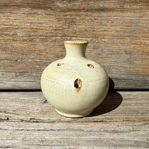 May include: A small, off-white ceramic vase with five holes around the top. The vase has a round body and a short neck.