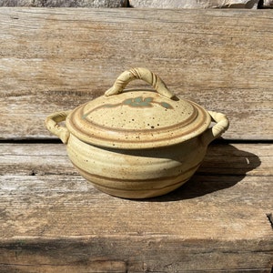 May include: A tan ceramic lidded bowl with a handle on the lid and two side handles. The lid has a leaf design and the bowl has a speckled appearance. The bowl is sitting on a weathered wooden surface.