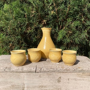 May include: A set of five ceramic sake cups and a pitcher in a pale yellow color. The cups and pitcher have a decorative design of stylized leaves.
