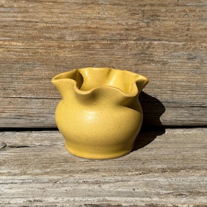 May include: A small, yellow ceramic vase with a ruffled rim. The vase has a rounded body and a speckled texture. The background is a weathered wooden surface.