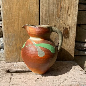 May include: A brown ceramic pitcher with a green and gray abstract design. The pitcher has a rounded body and a handle.