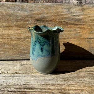 May include: A small, green and blue ceramic vase with a ruffled rim. The vase has a light gray base and a darker green glaze on the top.