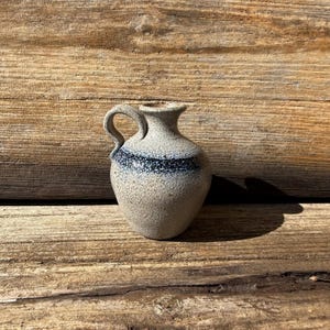 May include: A small, gray ceramic pitcher with a blue and white band around the top. The pitcher has a textured surface and a handle.