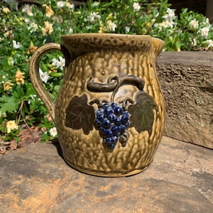 May include: A brown ceramic pitcher with a textured surface and a grapevine design. The pitcher has a handle and a spout. The grapes are blue and the leaves are green.