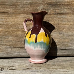 May include: A colorful ceramic pitcher with a handle. The top is dark brown, with yellow, green, and pink paint dripping down the sides. The pitcher is set against a wooden background.