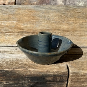 May include: A blue ceramic candle holder with a single candle holder in the center. The holder has a wide, shallow base and a raised center for the candle.