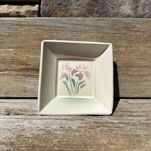 May include: A small, square, white ceramic dish with a raised rim. The dish has a floral design of pink and green irises.