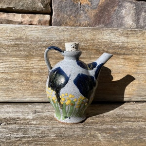 May include: A small, gray ceramic pitcher with a cork stopper. The pitcher has a blue and black floral design with yellow accents.