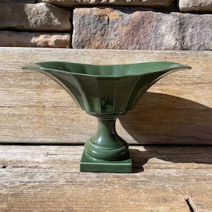 May include: A dark green ceramic pedestal bowl with a wide, flared rim and a fluted design. The bowl sits on a square base.