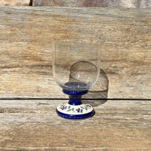 May include: A clear glass wine goblet with a blue and white floral design on the base. The goblet is sitting on a wooden surface.