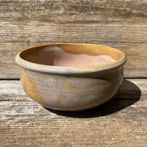 May include: A small, round ceramic bowl with a light brown glaze. The bowl has a subtle iridescent sheen and a slightly uneven surface.