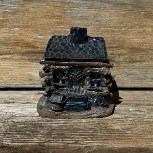 May include: A small, blue and brown ceramic cabin with a chimney and a barrel in front. The cabin has a log cabin style with a detailed front door and windows.