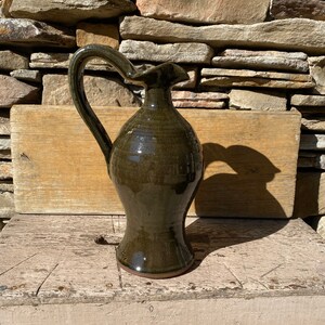 May include: A dark green ceramic pitcher with a wide mouth and a curved handle. The pitcher has a glossy finish and is sitting on a wooden surface.