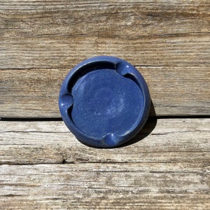 May include: A blue ceramic ashtray with three indentations for cigarettes. The ashtray is round and has a speckled glaze.