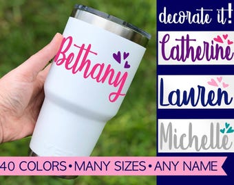 Puede incluir: Un vaso de acero inoxidable blanco con tapa transparente, con el nombre Bethany en letra cursiva rosa y tres corazones morados. Otros ejemplos incluyen Catherine, Lauren y Michelle con corazones. Texto: decorate it! 40 COLORS • MANY SIZES • ANY NAME.