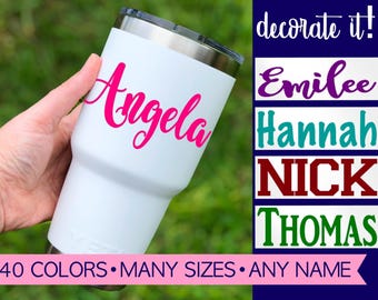 Puede incluir: Un vaso de acero inoxidable blanco con borde y tapa metálicos, que muestra el nombre Angela en letra cursiva rosa brillante. La imagen incluye el texto decorate it!, Emilee, Hannah, NICK, THOMAS y 40 COLORS MANY SIZES ANY NAME.
