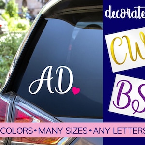May include: A custom vinyl decal for a car window featuring the letters AD in white script with a small pink heart. Additional examples show CW in gold with a red heart and BS in purple with a teal heart. Text reads decorate it!