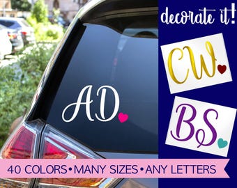 May include: A custom vinyl decal for a car window featuring the letters AD in white script with a small pink heart. Additional examples show CW in gold with a red heart and BS in purple with a teal heart. Text reads decorate it!