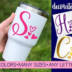 May include: White tumbler with a pink letter S and pink hearts. The tumbler is being held by a hand. The image also shows examples of other letters and colors. "Decorate it!" is written in the top right corner of the image. "40 COLORS. MANY SIZES. ANY LETTER" is written at the bottom of the image.