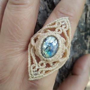 Puede incluir: Un anillo de macramé blanco con una piedra de vidrio azul iridiscente en el centro.