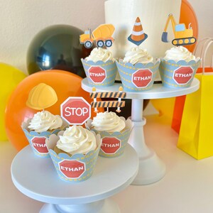 May include: Construction themed cupcakes with blue and yellow frosting. The cupcakes are decorated with construction signs, a stop sign, a hard hat, and a construction vehicle. The cupcakes are on a white cake stand and have the name "Ethan" written on them.