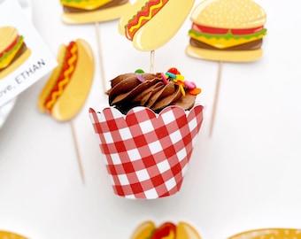 May include: A chocolate cupcake with red and white checkered cupcake liner and colorful sprinkles. The cupcake is topped with a hot dog, hamburger, and other food-themed decorations.
